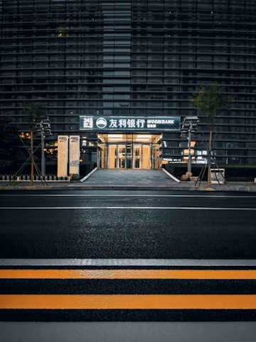 A modern building with a sleek outer facade consisting of horizontal lines and glass panels. The entrance is illuminated, displaying signage with both Korean and English text, indicating a bank. The scene includes a crosswalk with yellow safety stripes in the foreground and some greenery alongside the path leading to the entrance.