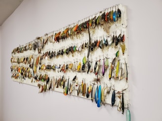 A collection of fishing lures is neatly organized on a white wall display board. The lures vary in color, size, and style, featuring a range of metallic hues and intricate designs. Each lure is equipped with hooks and displayed in an orderly manner.