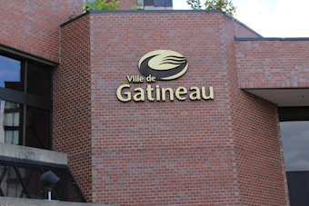 A brick building fa&ccedil;ade features a sign with the text 'Ville de Gatineau' accompanied by a logo. The sign is mounted on the building's wall, which is composed of red bricks. There are window reflections visible on the left side of the image.