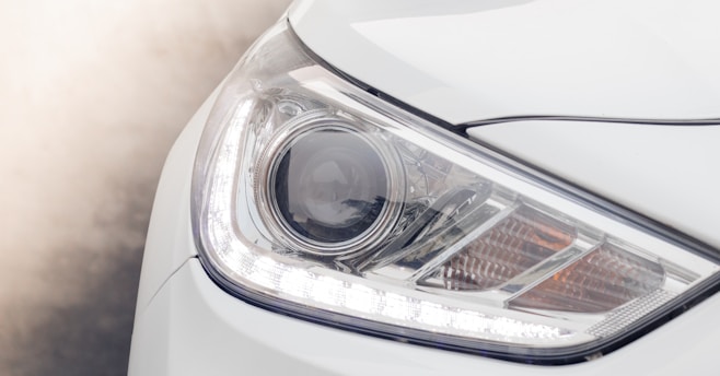 Close-up of a modern car headlight featuring a sleek, angular design with LED elements. The surrounding area is part of the vehicle’s white body exterior.
