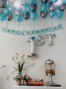 Blue and silver balloons are arranged on a ceiling with a banner that reads 'Happy Birthday.' The focus is on large silver foil balloons shaped like '1st.' Below, a table displays a decorative floral arrangement, a cake, a bottle of wine, and a drink dispenser.