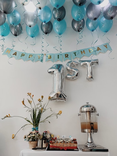 Blue and silver balloons are arranged on a ceiling with a banner that reads 'Happy Birthday.' The focus is on large silver foil balloons shaped like '1st.' Below, a table displays a decorative floral arrangement, a cake, a bottle of wine, and a drink dispenser.