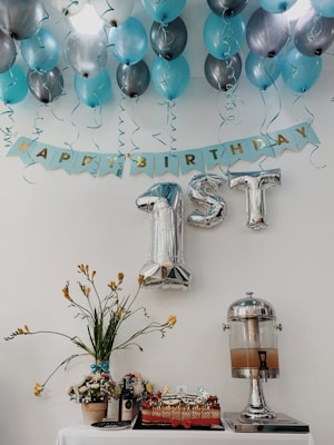 Blue and silver balloons are arranged on a ceiling with a banner that reads 'Happy Birthday.' The focus is on large silver foil balloons shaped like '1st.' Below, a table displays a decorative floral arrangement, a cake, a bottle of wine, and a drink dispenser.