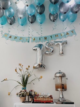Blue and silver balloons are arranged on a ceiling with a banner that reads 'Happy Birthday.' The focus is on large silver foil balloons shaped like '1st.' Below, a table displays a decorative floral arrangement, a cake, a bottle of wine, and a drink dispenser.