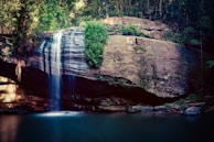 A serene waterfall cascades gently over a large, rugged rock formation surrounded by lush greenery. The water flows into a calm, reflective pool below, creating a tranquil natural scene.