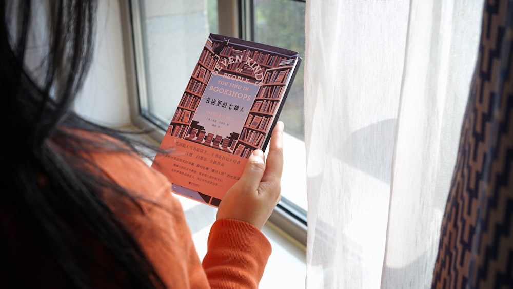 a woman holding up a book in front of a window