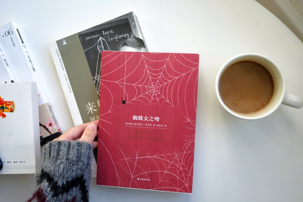 a person holding a book next to a cup of coffee