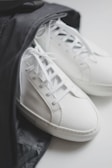 A pair of white leather sneakers partially inside a black bag. The shoes have white laces and a minimalistic design, highlighting a clean and modern look.