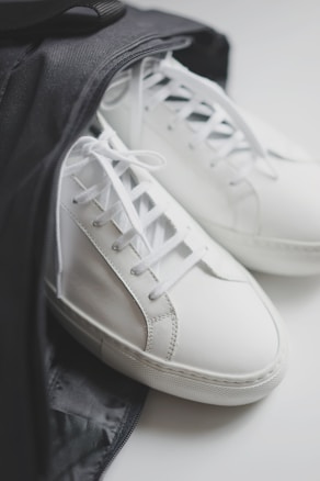 A pair of white leather sneakers partially inside a black bag. The shoes have white laces and a minimalistic design, highlighting a clean and modern look.