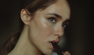A person with light skin and brown hair is biting a piece of dark chocolate. The lighting is soft, highlighting their facial features with a gentle shadow.