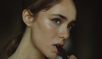 A person with light skin and brown hair is biting a piece of dark chocolate. The lighting is soft, highlighting their facial features with a gentle shadow.