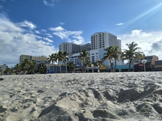 Sandy beach with palm trees leading up to a row of multi-story hotel buildings under a partly cloudy sky. The beach area appears quiet with soft sand, and there are various small structures and signs indicating businesses along the beachfront.