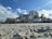 Sandy beach with palm trees leading up to a row of multi-story hotel buildings under a partly cloudy sky. The beach area appears quiet with soft sand, and there are various small structures and signs indicating businesses along the beachfront.