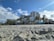 Sandy beach with palm trees leading up to a row of multi-story hotel buildings under a partly cloudy sky. The beach area appears quiet with soft sand, and there are various small structures and signs indicating businesses along the beachfront.