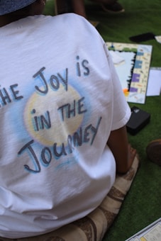 A person is sitting on a cushion wearing a white t-shirt with the text 'The Joy is in the Journey' printed on it. Nearby, there is a board game with various cards and pieces scattered on the grass.