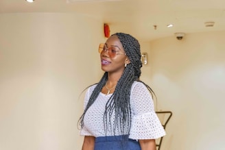 A woman with long braided hair wearing sunglasses and a white, lacy top. She seems to be smiling gently and wearing a blue skirt. The background is a plain, light-colored wall with some ceiling fixtures visible.