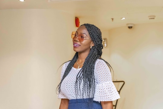 A woman with long braided hair wearing sunglasses and a white, lacy top. She seems to be smiling gently and wearing a blue skirt. The background is a plain, light-colored wall with some ceiling fixtures visible.