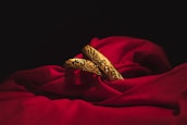Two intricately designed gold bangles rest elegantly on a rich, red fabric. The deep shadows of the surrounding area contrast with the vibrant brightness of the gold and the fabric, highlighting the detailed floral patterns etched into the metal.