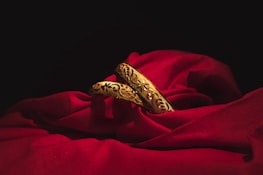 Two intricately designed gold bangles rest elegantly on a rich, red fabric. The deep shadows of the surrounding area contrast with the vibrant brightness of the gold and the fabric, highlighting the detailed floral patterns etched into the metal.