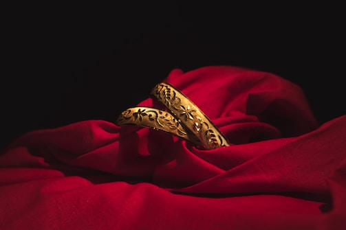 Two intricately designed gold bangles rest elegantly on a rich, red fabric. The deep shadows of the surrounding area contrast with the vibrant brightness of the gold and the fabric, highlighting the detailed floral patterns etched into the metal.