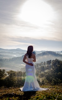 A woman dressed in sage linen layers standing on a sunlit mountain overlook with a gentle breeze.