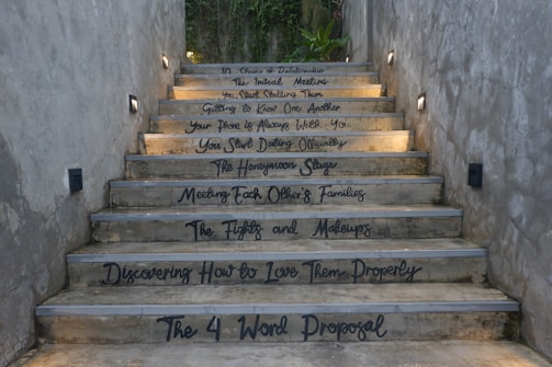 A staircase with motivational and relationship-themed phrases written on each step. The setting is outdoors, surrounded by concrete walls and minimal lighting. At the top, there is greenery visible.