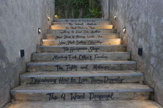 A staircase with motivational and relationship-themed phrases written on each step. The setting is outdoors, surrounded by concrete walls and minimal lighting. At the top, there is greenery visible.