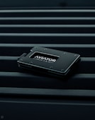 A sleek, minimalist wallet labeled 'AVIATOR' rests on a surface with parallel lines. The wallet is dark-colored with metallic accents, conveying a modern and stylish design.