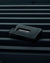 A sleek, minimalist wallet labeled 'AVIATOR' rests on a surface with parallel lines. The wallet is dark-colored with metallic accents, conveying a modern and stylish design.