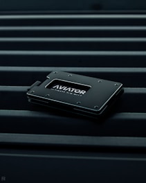 A sleek, minimalist wallet labeled 'AVIATOR' rests on a surface with parallel lines. The wallet is dark-colored with metallic accents, conveying a modern and stylish design.
