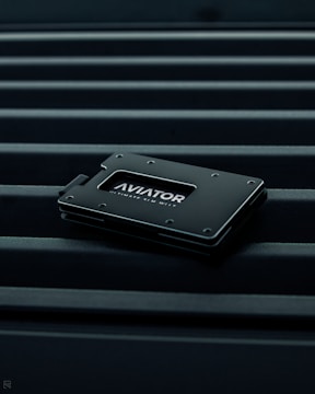A sleek, minimalist wallet labeled 'AVIATOR' rests on a surface with parallel lines. The wallet is dark-colored with metallic accents, conveying a modern and stylish design.