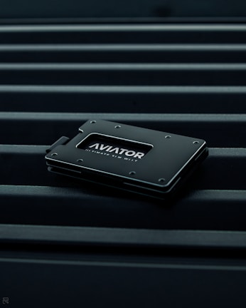 A sleek, minimalist wallet labeled 'AVIATOR' rests on a surface with parallel lines. The wallet is dark-colored with metallic accents, conveying a modern and stylish design.