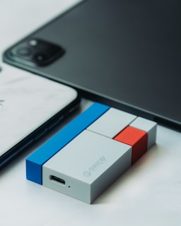 A close-up view of a USB device with a modular design, featuring colored blocks in blue, white, red, and gray. The device is placed on a surface next to a smartphone and part of a tablet.
