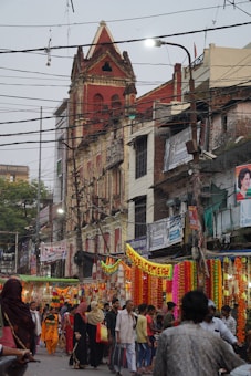 A busy street scene featuring a crowd of people walking through a vibrant outdoor market. Colorful garlands and decorations are hung overhead, adding a festive atmosphere. The surrounding architecture includes a prominent red-brick building with intricate designs and several stories. Numerous wires crisscross the scene above the street.