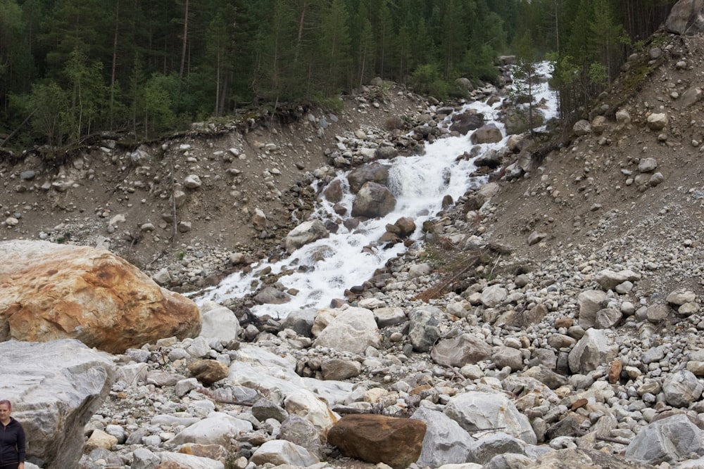 a man standing on a rocky hillside next to a waterfall