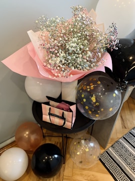 A bouquet of pink and white baby's breath flowers wrapped in pink paper, placed on a small black table. Surrounding the bouquet are several balloons in shades of white, black, and gold with transparent sections. There's also a pink and black striped shopping bag on the table.