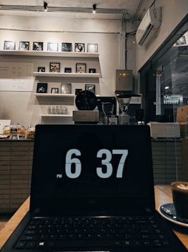 A dimly lit coffee shop interior featuring a laptop with a time display of 6:37 PM in the foreground. Behind the laptop, there are shelves with multiple framed pictures and artworks, a menu on the wall, and various coffee-making equipment such as an espresso machine and grinders. A cup of coffee rests on the table next to the laptop.