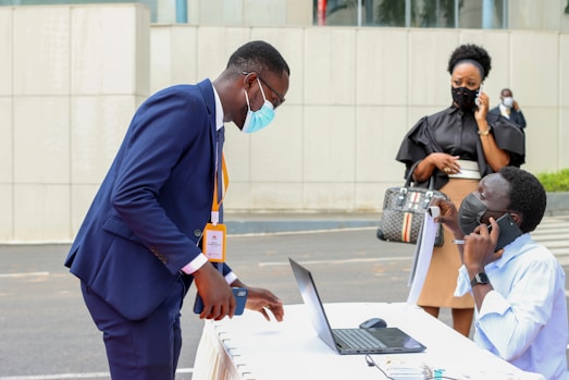 A person in a dark blue suit bends over a table with a laptop, interacting with another person seated and talking on a phone. They are both wearing masks. In the background, a woman dressed in black is also on the phone, standing nearby. The setting appears to be outdoor with an urban backdrop.