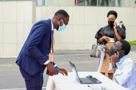 A person in a dark blue suit bends over a table with a laptop, interacting with another person seated and talking on a phone. They are both wearing masks. In the background, a woman dressed in black is also on the phone, standing nearby. The setting appears to be outdoor with an urban backdrop.