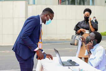 A person in a dark blue suit bends over a table with a laptop, interacting with another person seated and talking on a phone. They are both wearing masks. In the background, a woman dressed in black is also on the phone, standing nearby. The setting appears to be outdoor with an urban backdrop.