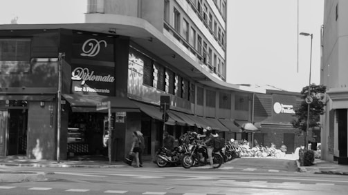 The image depicts a street scene with several motorcycles parked along the sidewalk in front of a restaurant named Diplomata. The restaurant has a black awning and a sign with its name. There are pedestrians walking on the sidewalk and crossing the street. In the background, there are other buildings and a sign for Banco Paulista.