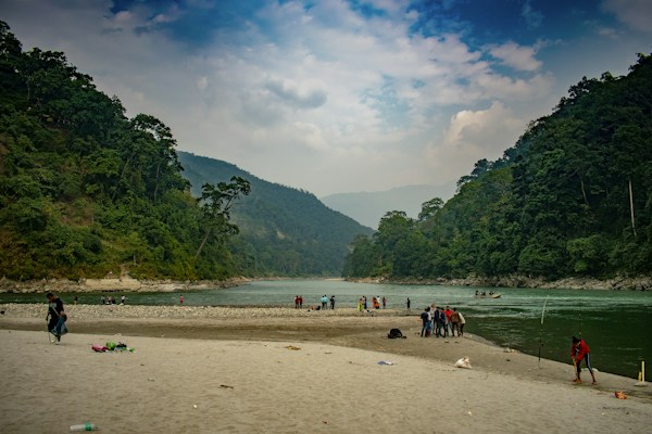A serene landscape with a river flowing gently between lush green hills under a partly cloudy sky. People are gathered by the riverbank, engaging in activities such as fishing and walking. The sandy shores and dense foliage create a picturesque scene of natural beauty.