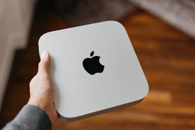 A hand is holding a small, sleek, square-shaped electronic device with a prominent black Apple logo on its silver surface. The background is blurred and shows wooden flooring.