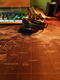 A black fitness tracker is placed on top of a world map-themed desk mat. In the background, a mechanical keyboard with RGB lighting is visible, displaying various colors. There's a cable running alongside the desk, and some books or folders are stacked on the side, partially shown.