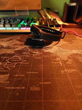 A black fitness tracker is placed on top of a world map-themed desk mat. In the background, a mechanical keyboard with RGB lighting is visible, displaying various colors. There's a cable running alongside the desk, and some books or folders are stacked on the side, partially shown.