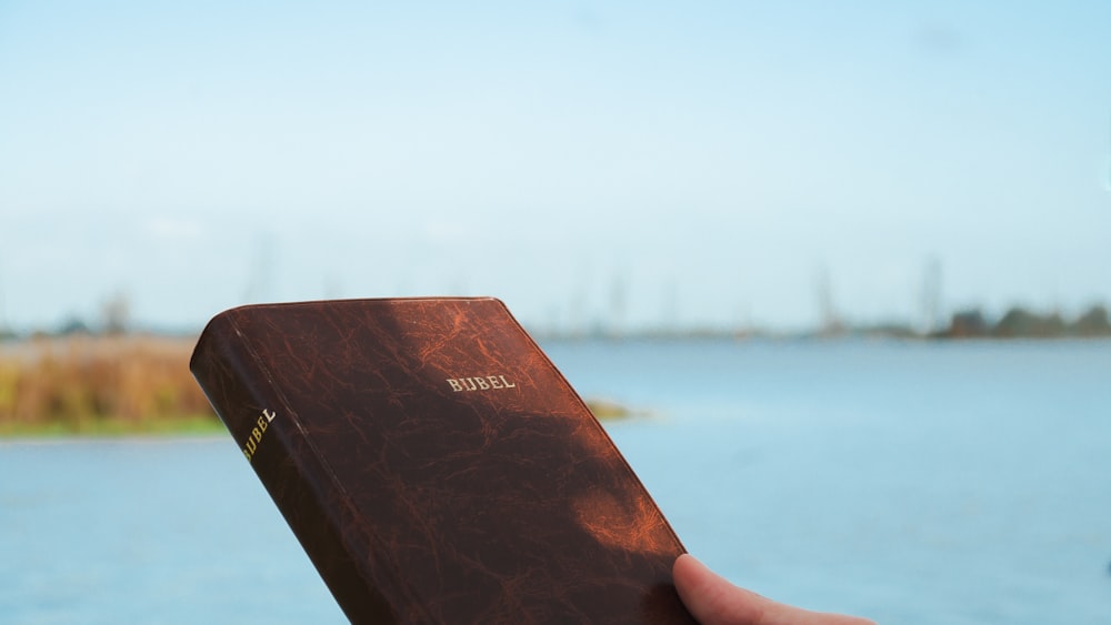 a hand holding a brown book over a body of water