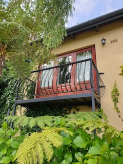 A small balcony with decorative black railings is attached to a house with light yellow walls. The balcony doors are made of dark wood with glass panels, partially covered by white curtains. Surrounding the structure are lush green plants and ferns with a variety of leaves and textures. A small exterior light fixture is mounted on the wall near the balcony.