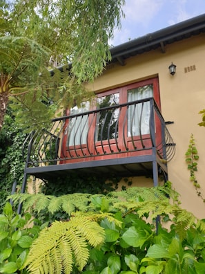 A small balcony with decorative black railings is attached to a house with light yellow walls. The balcony doors are made of dark wood with glass panels, partially covered by white curtains. Surrounding the structure are lush green plants and ferns with a variety of leaves and textures. A small exterior light fixture is mounted on the wall near the balcony.