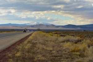 A sprawling landscape with a road leading toward distant mountains under a sky filled with clouds. Several vehicles are visible on the road, suggesting travel or a journey. The foreground consists of dry grass and shrubs, typical of a semi-arid region.