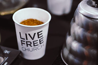 A coffee mug with the words 'terrible meeting coffee club' in gold lettering, sitting on a worn church basement table next to a folding chair.